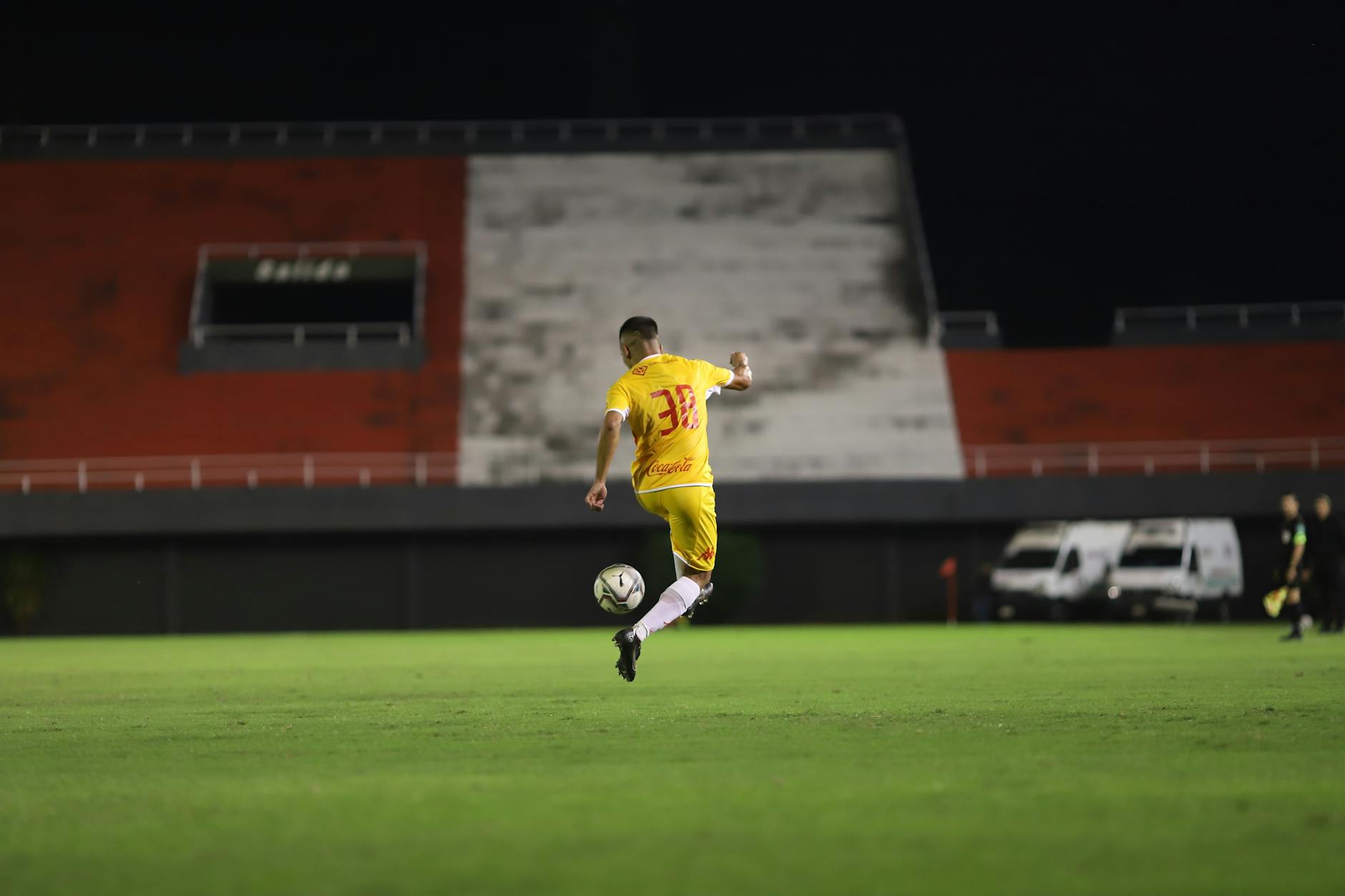 Soccer player in yellow jersey kicking a ball on a green pitch during night match under lights.